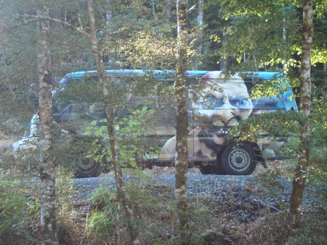 Paisaje boscoso con árboles en primer plano. Detrás se distingue una furgoneta con un rostro muy grande de Gollum pintado.;