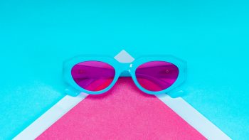 Gafas celestes con cristales de color rosa en el vértice de un triángulo rosa con borde blanco, sobre un fondo de color celeste.