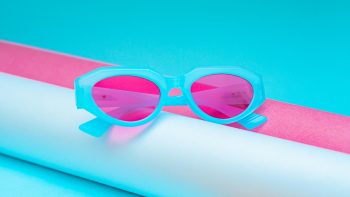 Gafas celestes con cristales de color rosa sobre dos tubos: uno blanco y otro rosa.